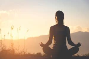 silhouette of a woman sitting in lotus position in front of setting sun wondering how can meditation help with addiction