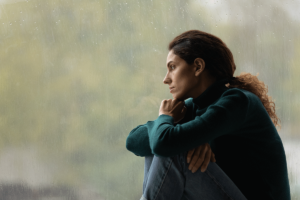 young dark-haired woman seated in front of large window on a rainy day struggling with the long-term effects of heroin addiction