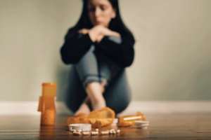 young woman sitting on floor staring at a collection of pill bottles wondering how can prescription drug addiction be treated.