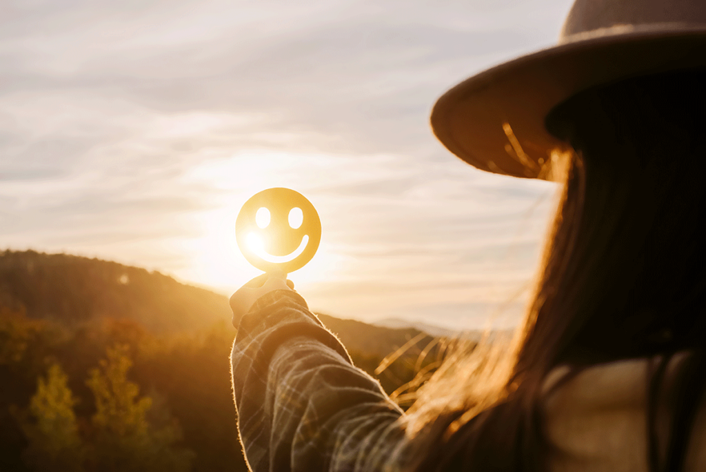 person outdoors at dawn holding up a smiley face cut out so that the rays of dawn shine through it as they finally understand what are the benefits of depression treatment