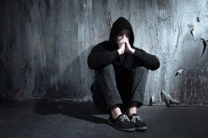 young man sitting on floor in dimly lit room with hood pulled over his head wondering what are the signs of a bipolar disorder