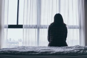 woman seated alone in her room on her bed staring out the window experiencing the long-term effects of heroin addiction