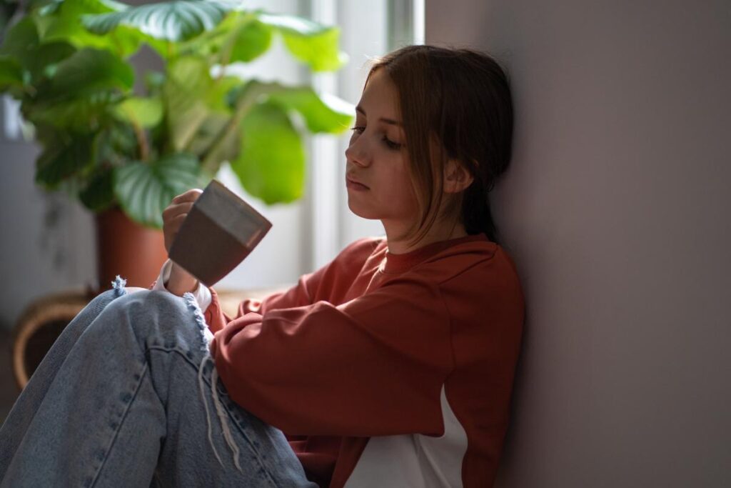 young girl seated listlessly against wall in front of window showing beautiful day outside and showing signs of prescription drug addiction.