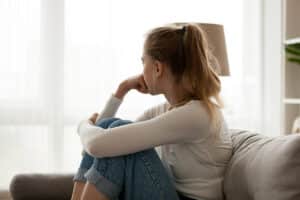 young woman seated alone on couch with her knees drawn up to her chest wondering how to calm social anxiety